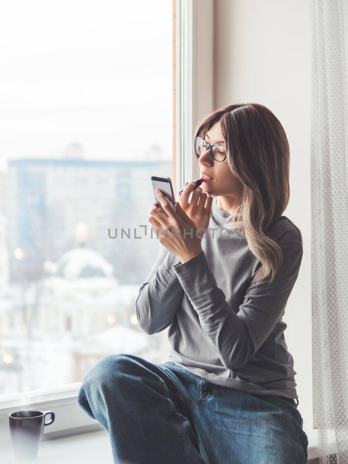 Woman with eyeglasses and curly hair paints her lips with lipstick and uses smartphone as mirror. Fast make-up. Morning routine. Cup of hot coffee on windowsill.