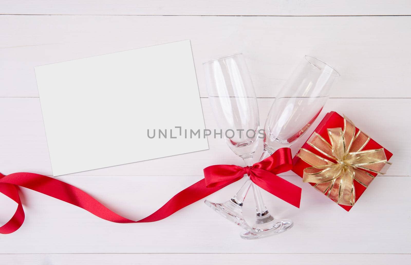 Valentine day concept, wineglass and red ribbon and gift box with greeting card on white wooden table background, champaign glass and postcard, top view, couples wine glass together, holiday concept.