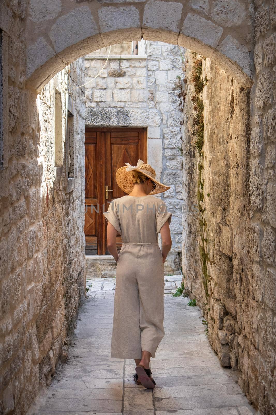 Rear view of beautiful blonde young female traveler wearing straw sun hat sightseeing and enjoying summer vacation in an old traditional costal town at Adriatic cost, Croatia by kasto