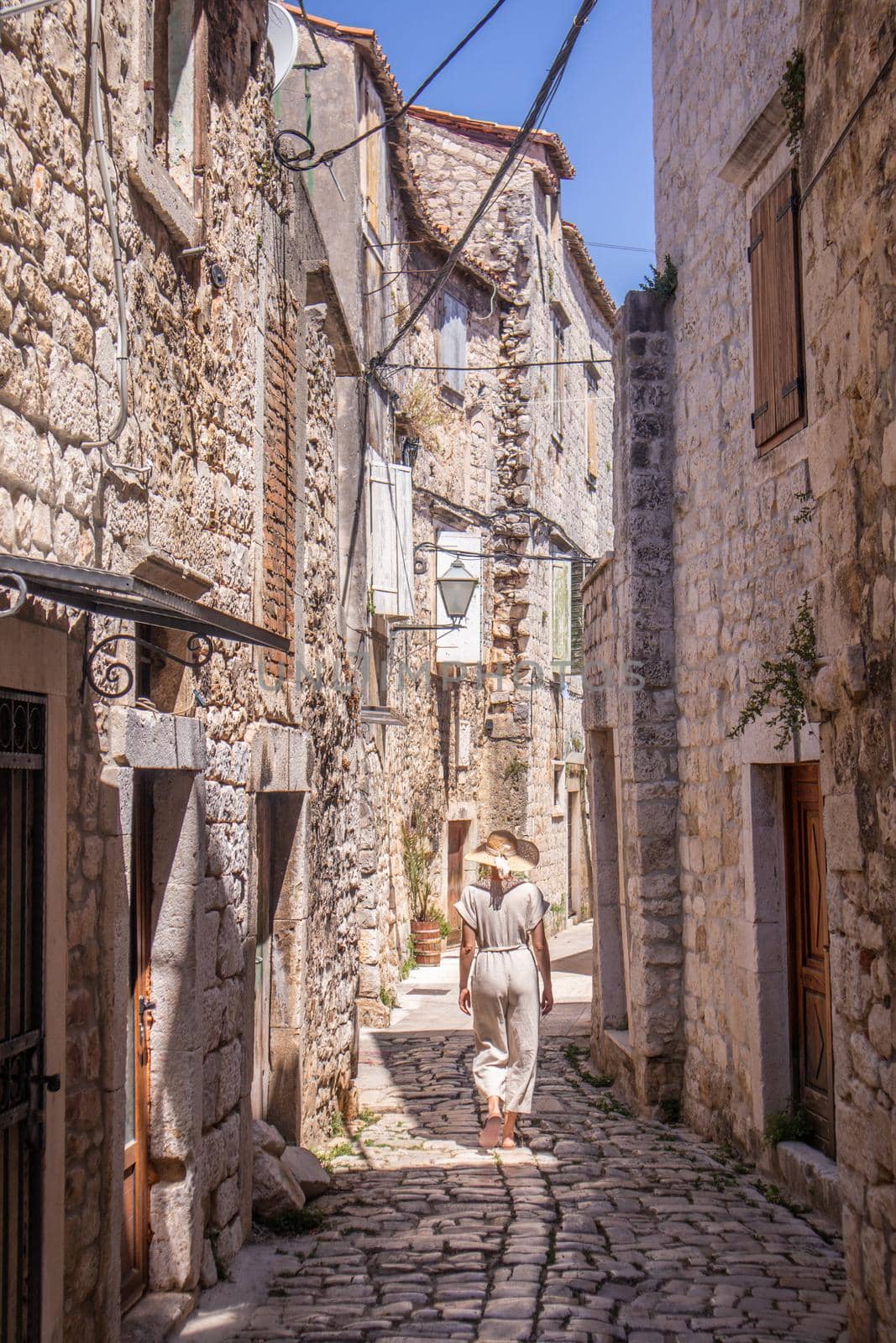 Rear view of beautiful blonde young female traveler wearing straw sun hat sightseeing and enjoying summer vacation in an old traditional costal town at Adriatic cost, Croatia.