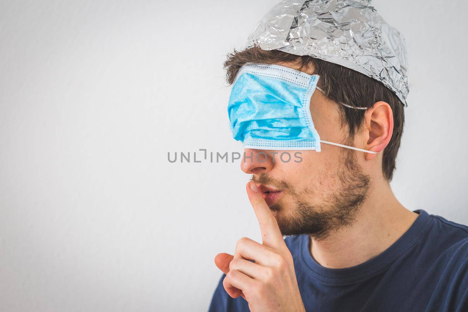 Conspiracy theory concept: Young man with face mask over the eyes and aluminum hat is making a psst! gesture by Daxenbichler