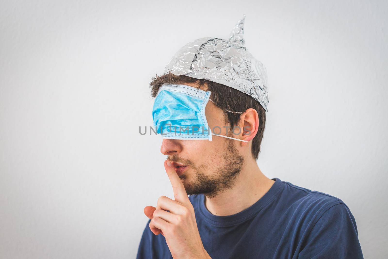 Conspiracy theory concept: Young man with face mask over the eyes and aluminum hat is making a psst! gesture by Daxenbichler