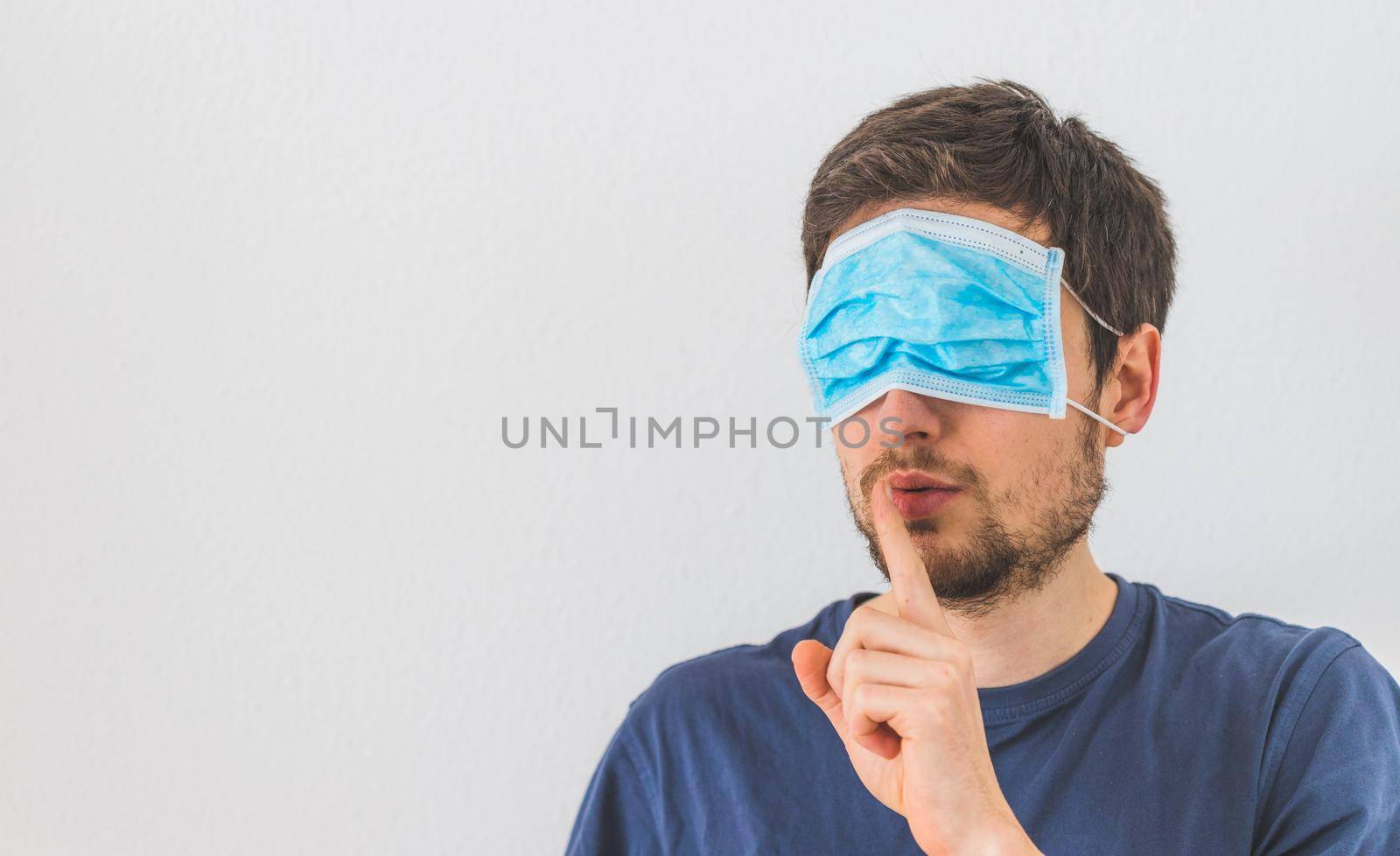 Conspiracy theory concept: Young man with face mask over the eyes is making a psst! gesture by Daxenbichler