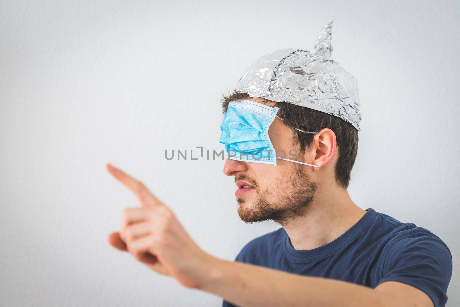 Conspiracy theory concept: Young angry man wearing aluminum hat and face mask over the eyes is gesturing angry. by Daxenbichler