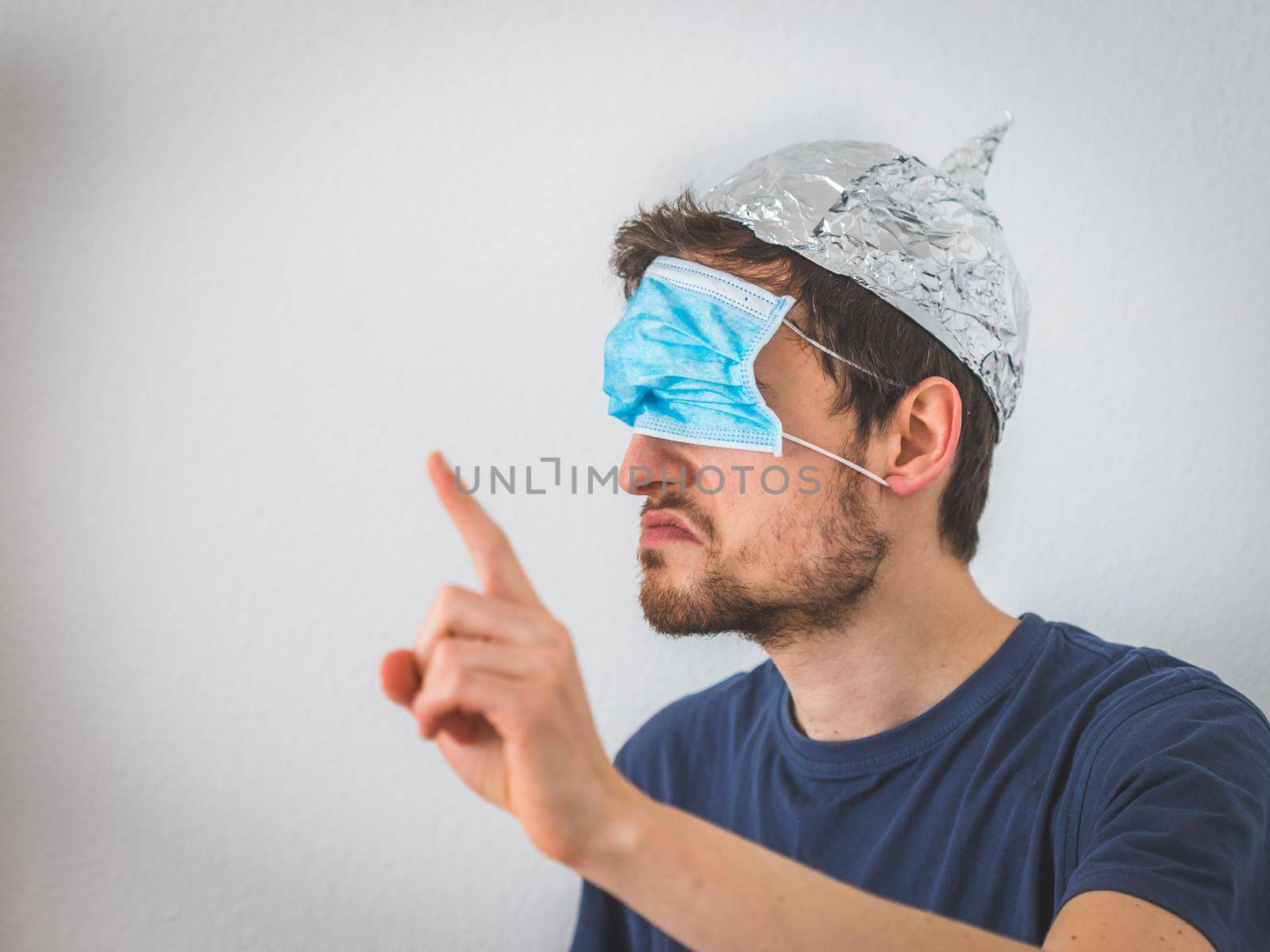 Conspiracy theory concept: Young angry man wearing aluminum hat and face mask over the eyes is gesturing angry. by Daxenbichler