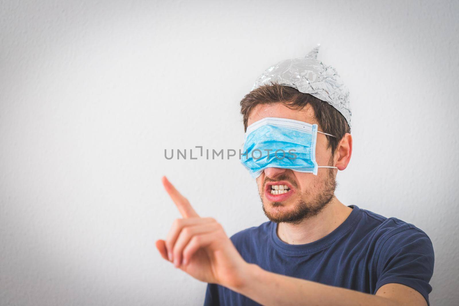 Conspiracy theory concept: Young angry man wearing aluminum hat and face mask over the eyes is gesturing angry. by Daxenbichler