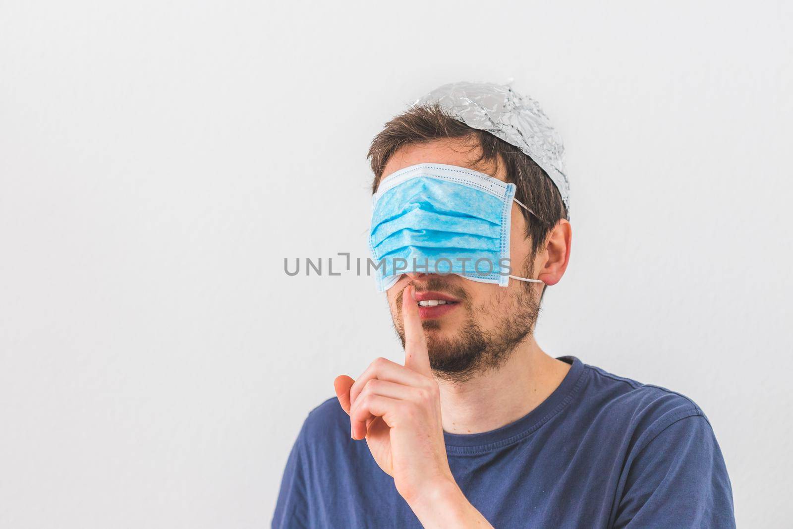 Conspiracy theory concept: Young man with face mask over the eyes and aluminum hat is making a psst! gesture by Daxenbichler