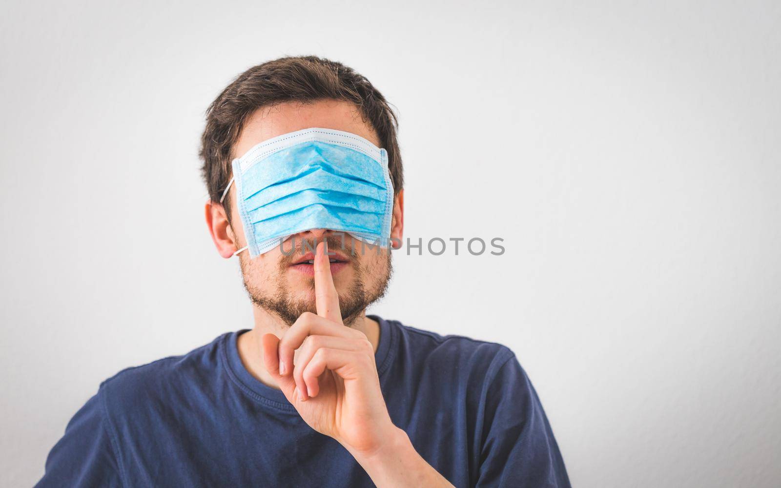 Conspiracy theory concept: Young man with face mask over the eyes is making a psst! gesture by Daxenbichler