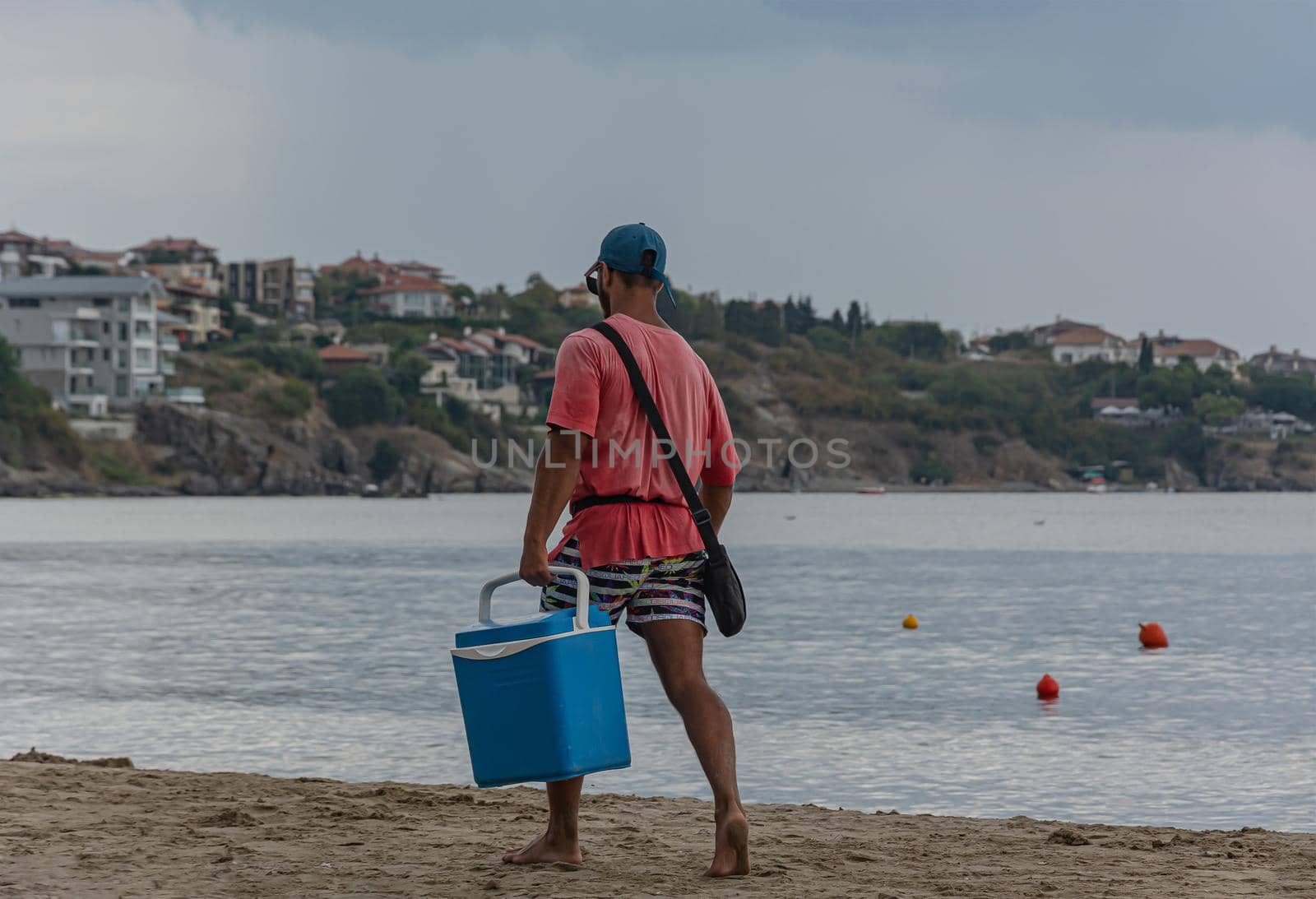 Sozopol, Bulgaria - 09/06/2018: a man in a red t-shirt with a blue box walks along the beach by Grommik