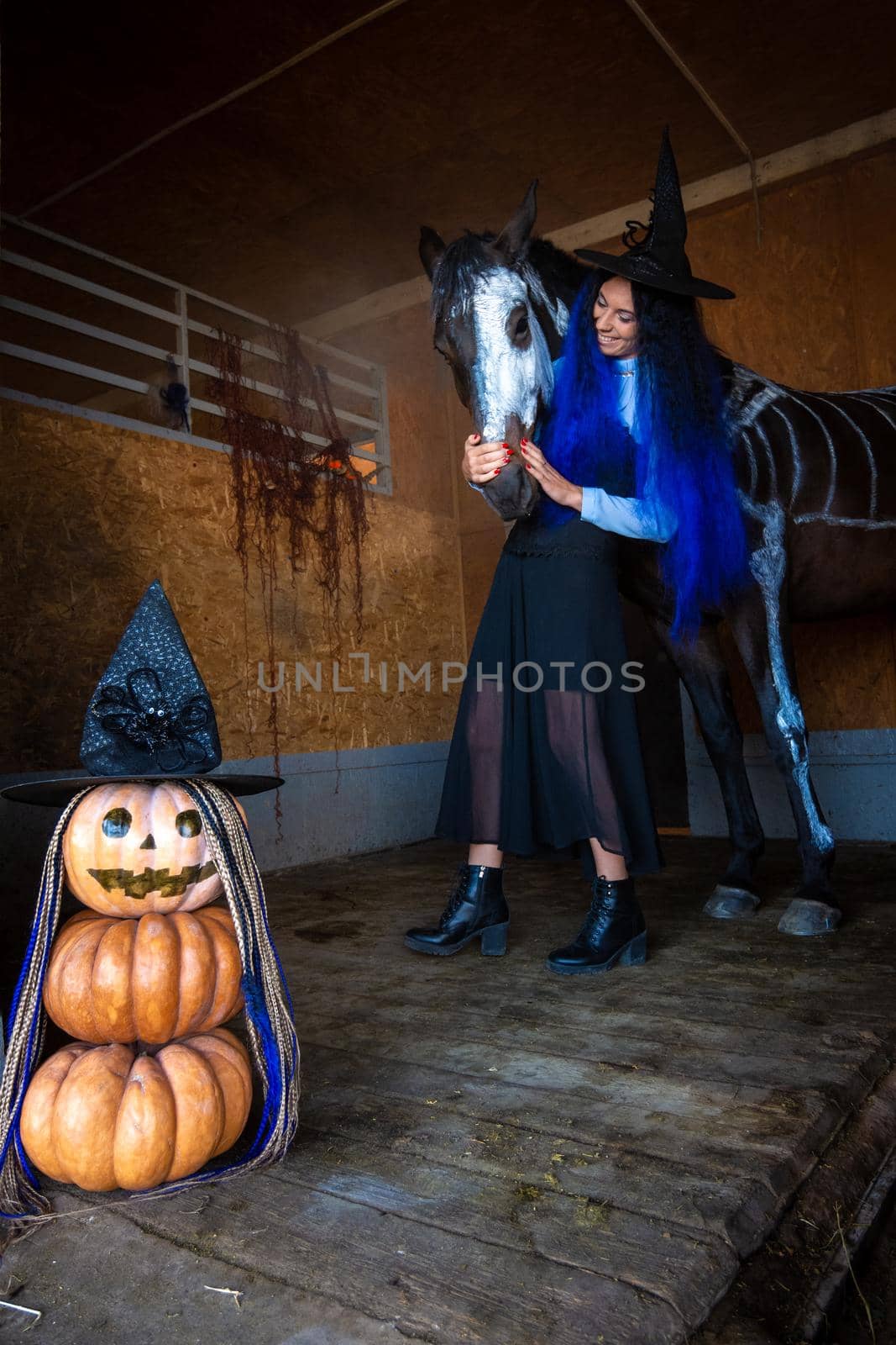 A girl in a witch costume hugs a horse in a corral, in the foreground an evil figure of pumpkins