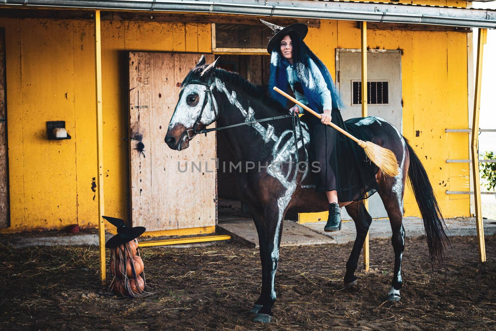 A girl dressed as a witch with a broom sits on a horse on which a skeleton is drawn by Madhourse