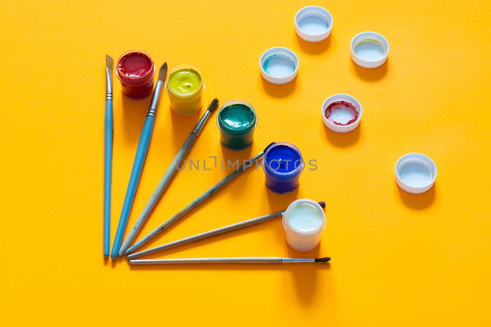 On a yellow background, brushes and jars of gouache are laid out, lids are randomly laid out