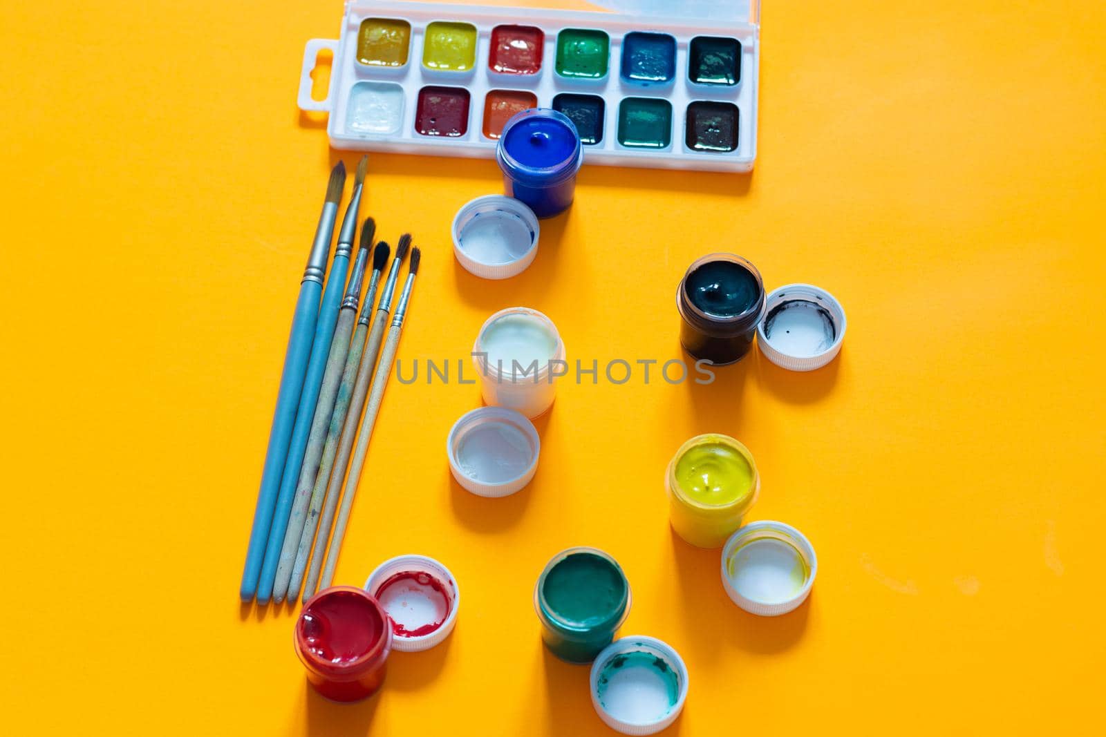 Brushes, jars of gouache and watercolor paints lie on a yellow background
