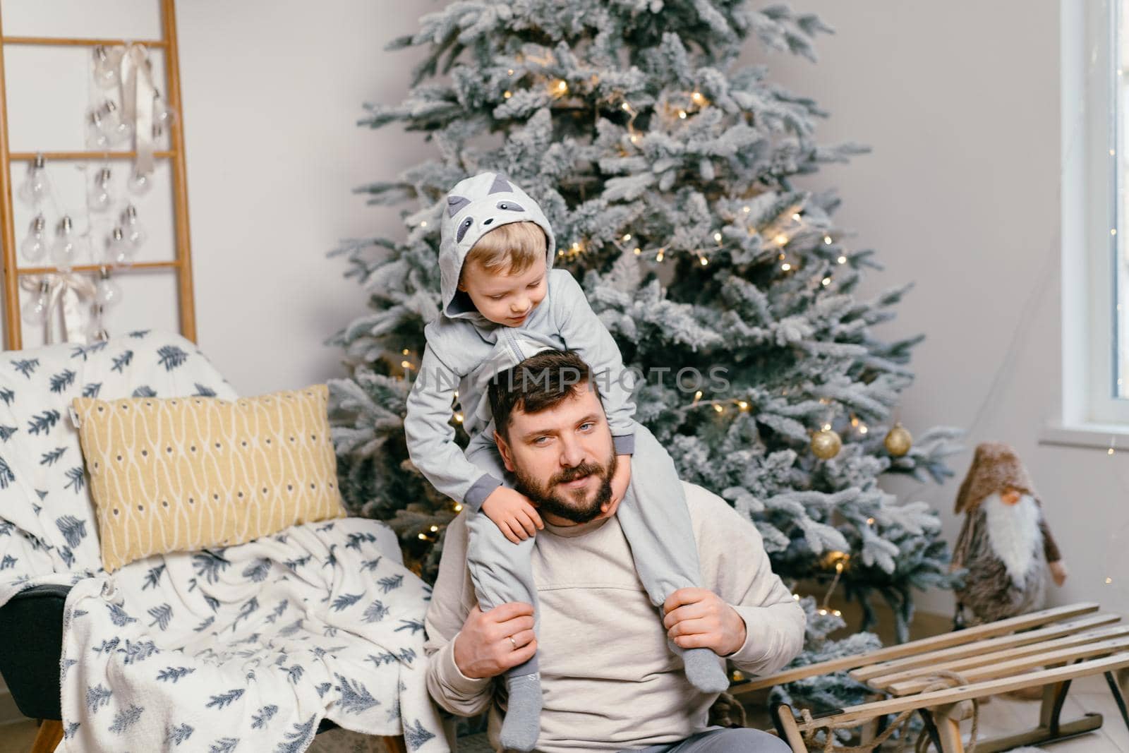 Holiday Christmas Handsome father playing with small cute son near decorated New year tree at home Family tradition European dad and his child son sitting on dad shoulders