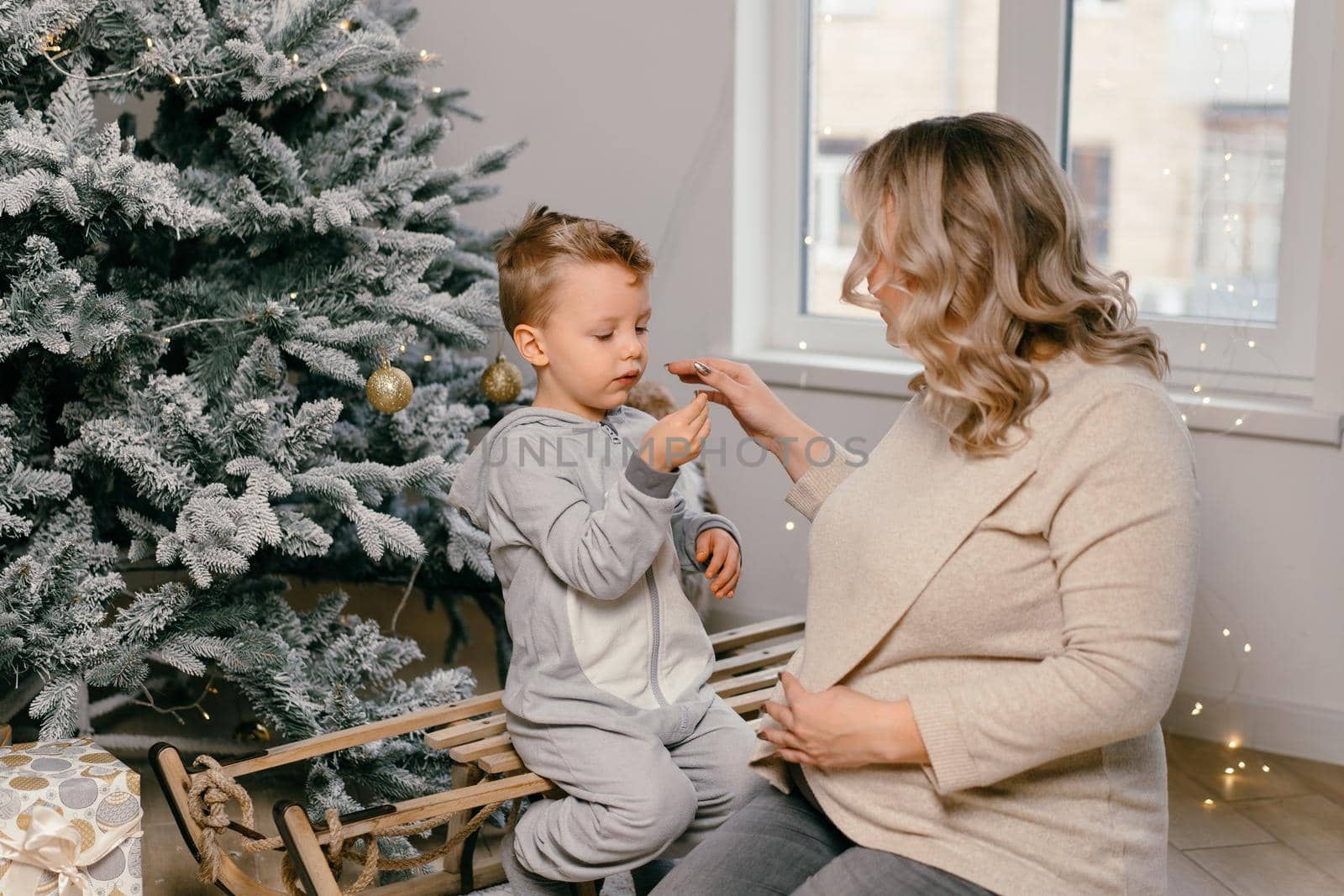Holiday Christmas Caucasian mother playing with little cute son near decorated New year tree at home Family tradition