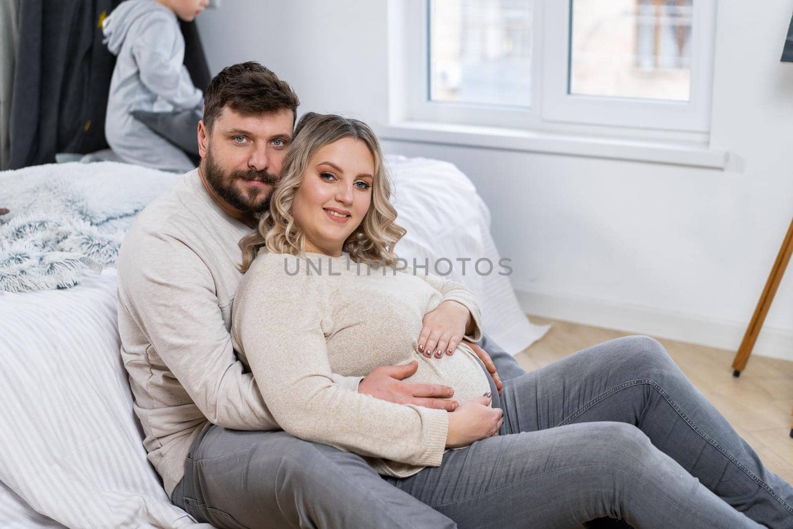 Happy family concept. Husband hug belly pregnant wife sitting floor indoor living room near sofa by andreonegin