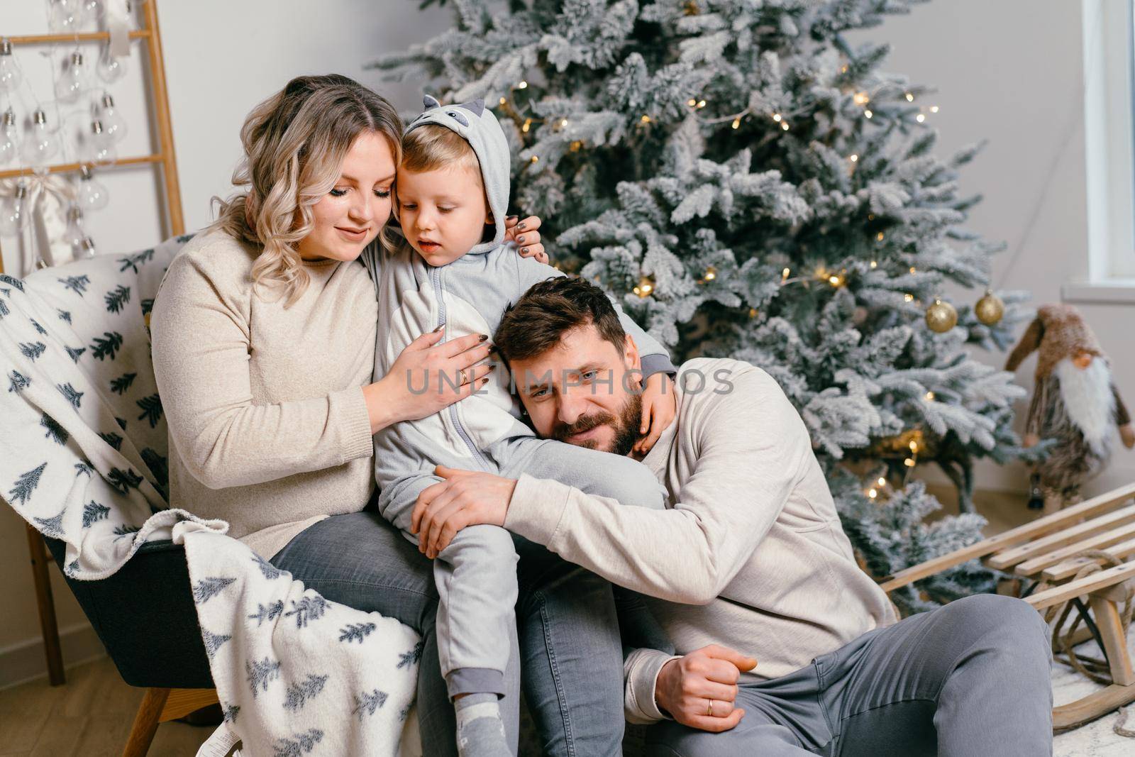 Christmas Family Happiness Portrait of dad, pregnant mom and little son sitting armchair at home near Christmas tree hug smile European young adult family holiday morning