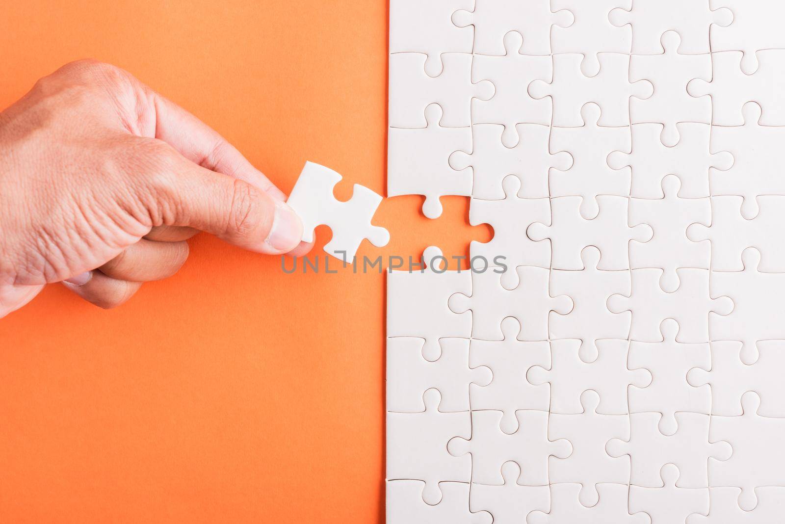 Top view flat lay of hand-holding last piece white paper jigsaw puzzle game last pieces put to place for solve problem complete mission, studio shot on an orange background, quiz calculation concept
