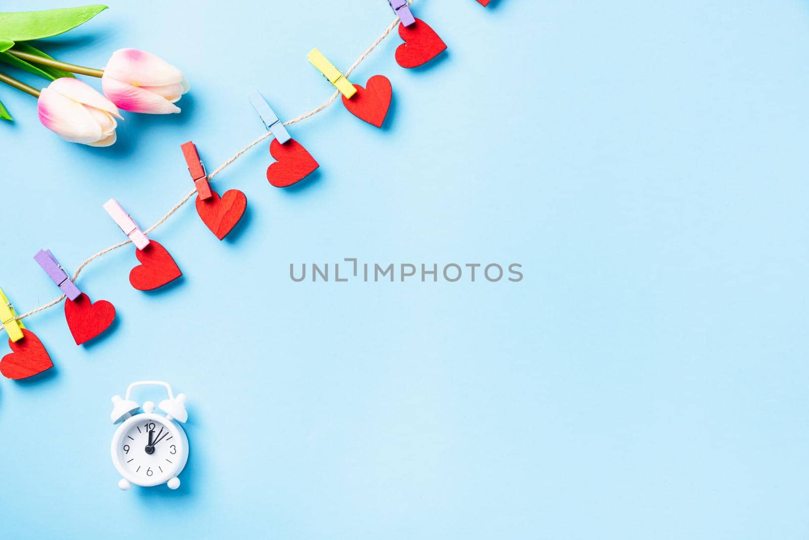 Happy Valentine's day concept. red heart-shaped valentines decoration hanging with clips on the rope for love and white alarm clock greeting card on blue background with copy space