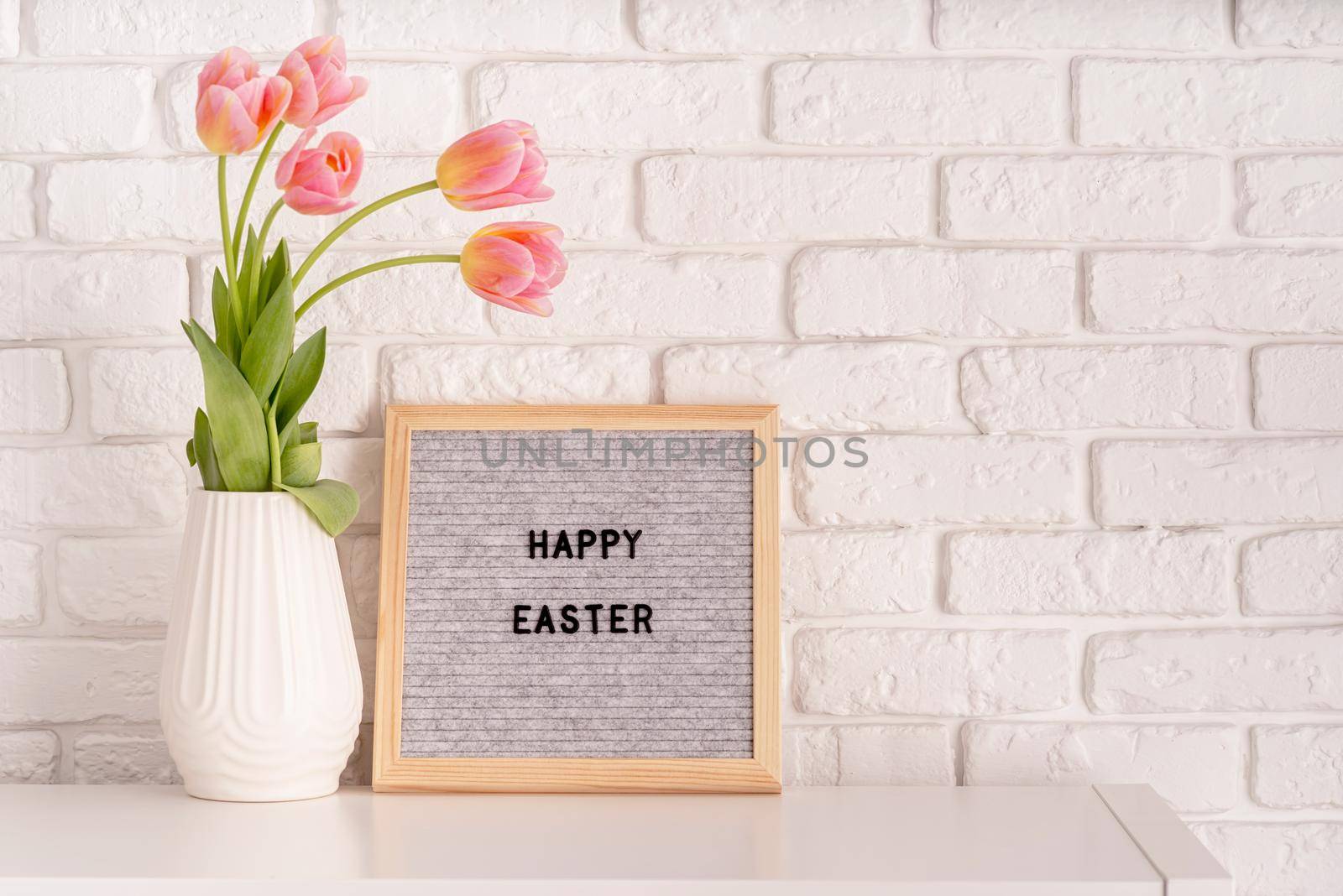 Easter hliday concept. Vase with tulips and gray felt letter board with words Happy Easter on white bricks background