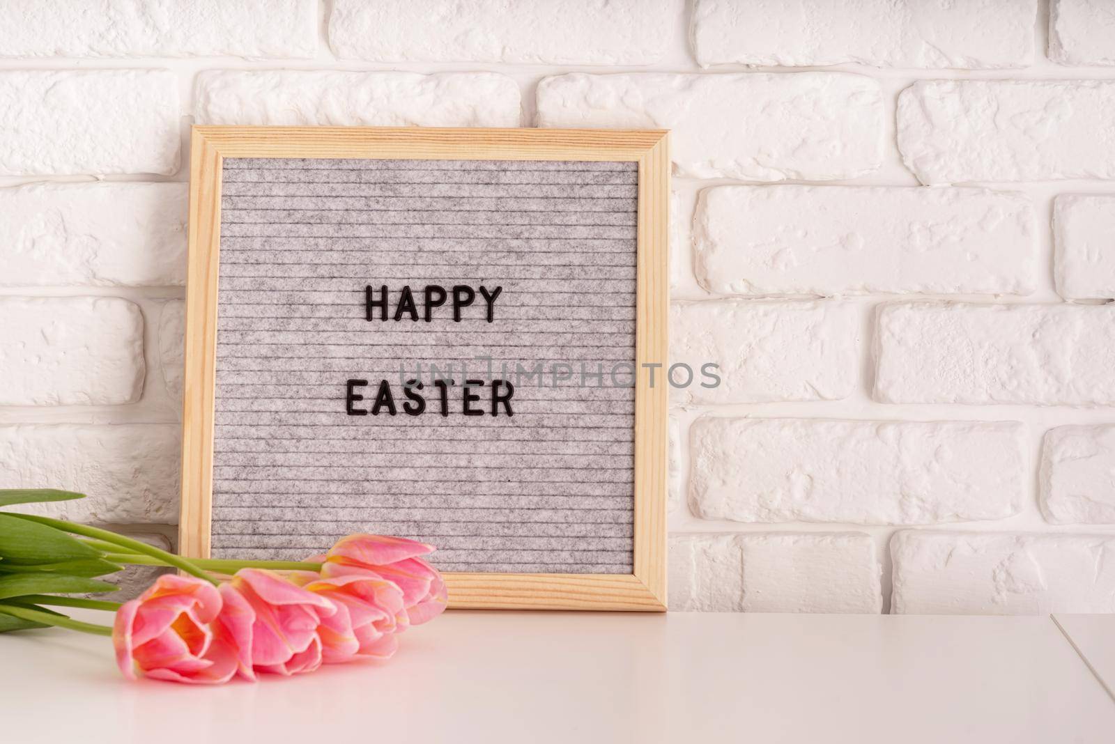 Easter hliday concept. Vase with tulips and gray felt letter board with words Happy Easter on white bricks background