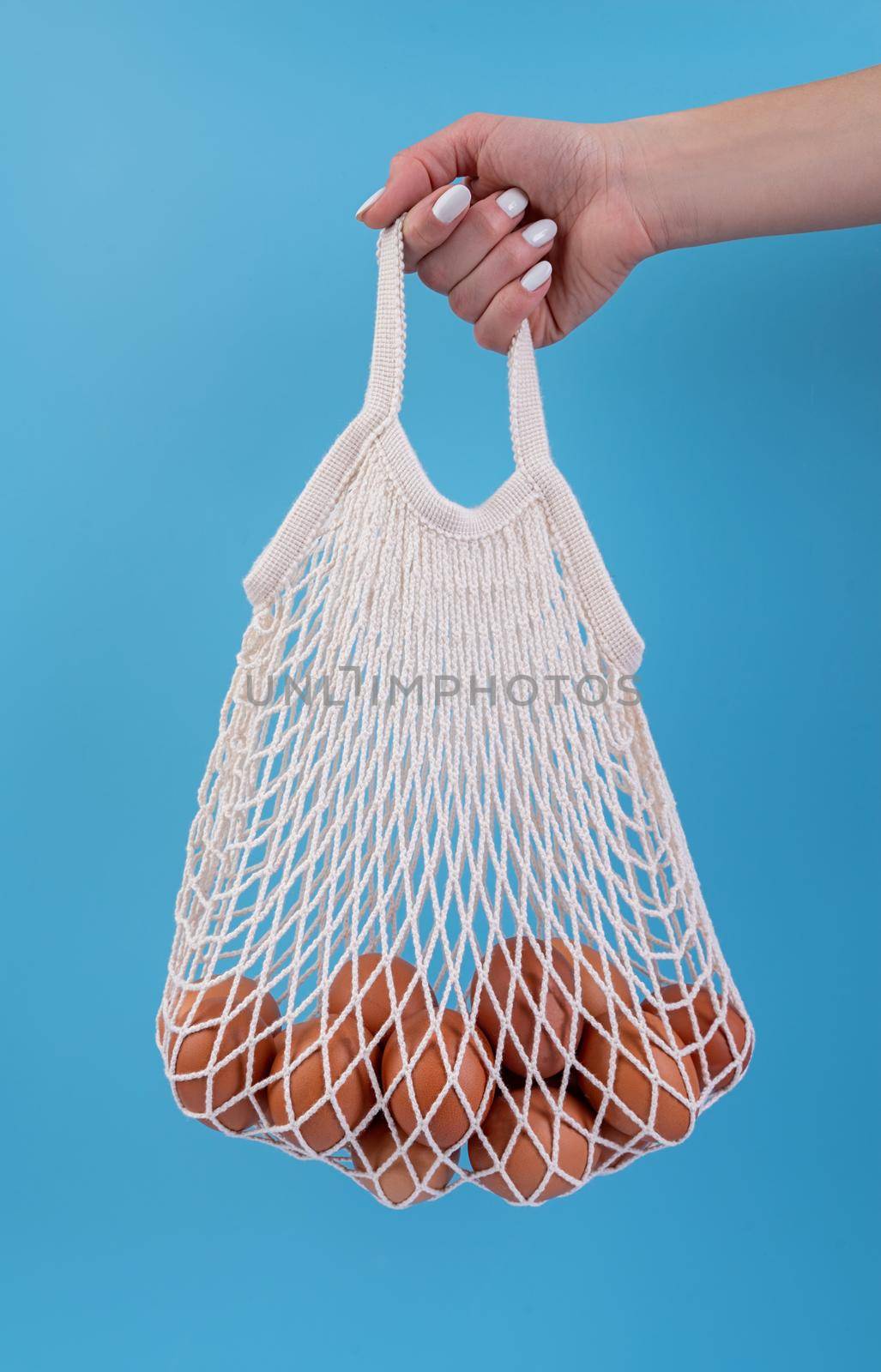 Woman hand holding eggs in a white shopping net bag isolated on blue background top view flat lay with copy space by Desperada
