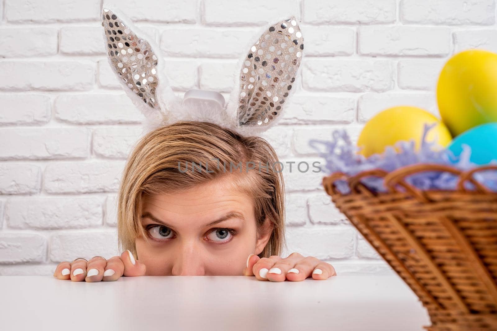 Easter holiday concept. Blond woman with bunny ears looking at the basket with Easter colored eggs on white brick background