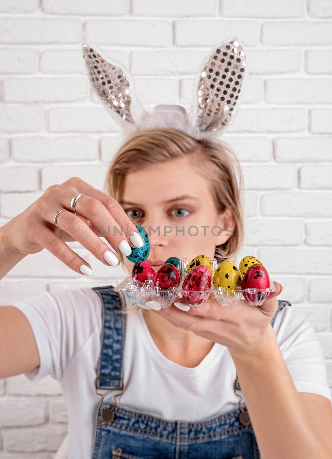 Young funny woman in bunny ears putting a colored quail egg into the plastic holder by Desperada