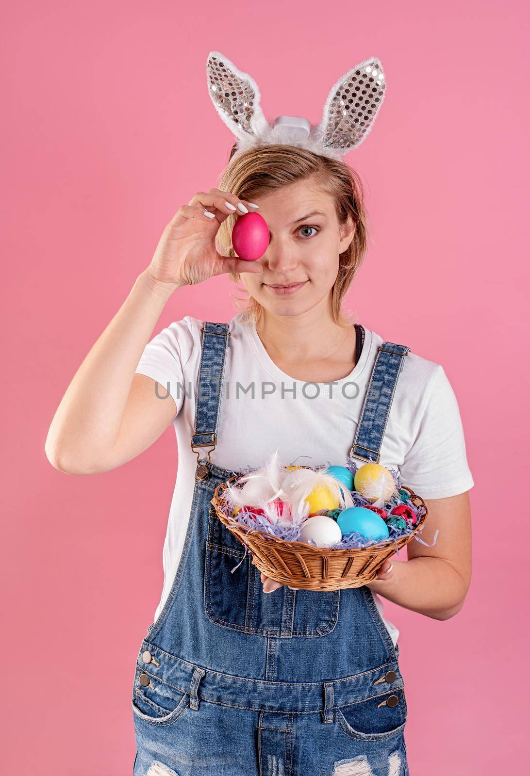 Young woman in bunny ears holding colored chicken eggs in front of her eye isolated on pink background by Desperada