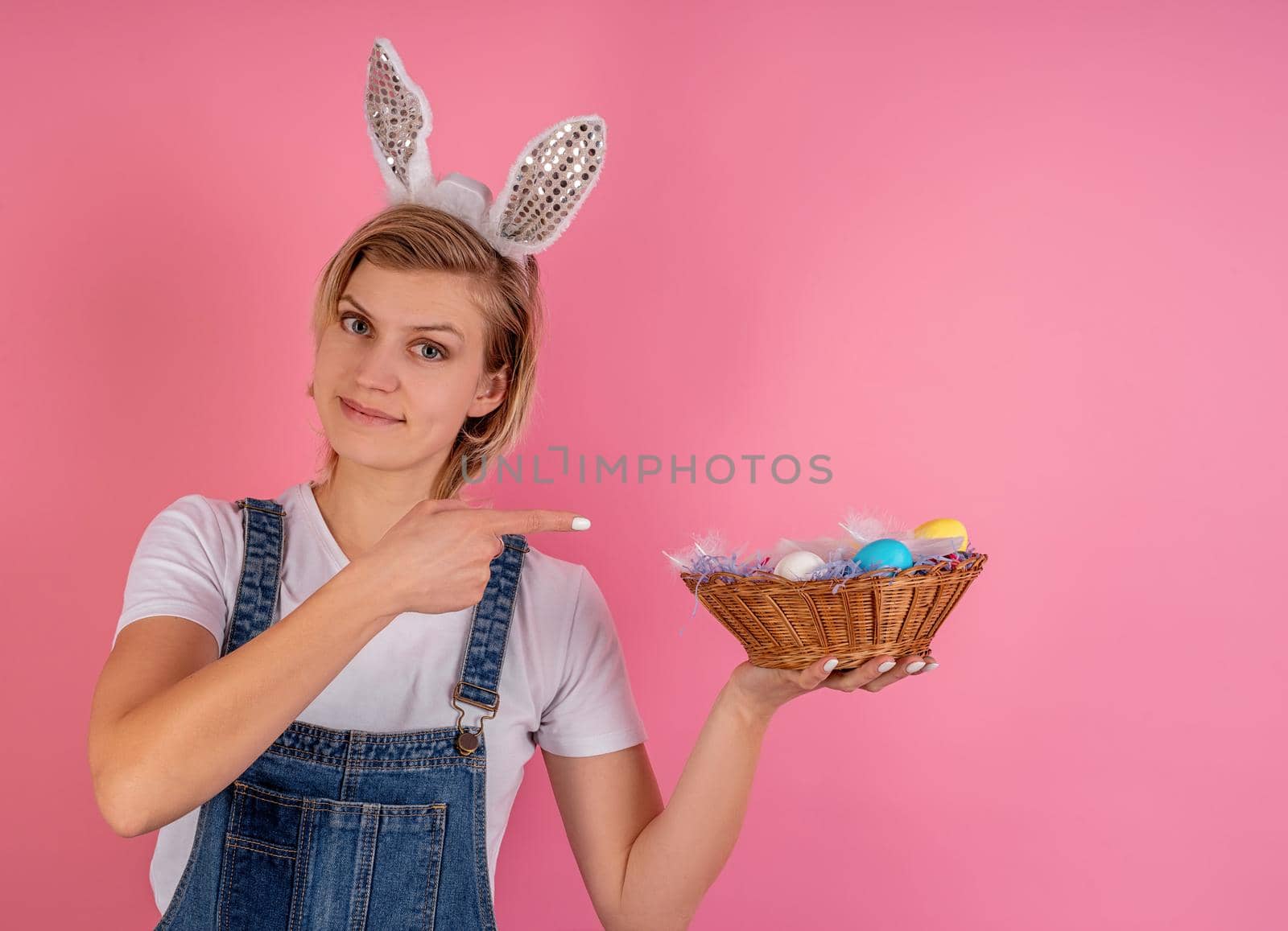Easter holiday concept. Young blond woman in bunny ears pointing to the basket with colored eggs and winking isolated on pink background with copy space