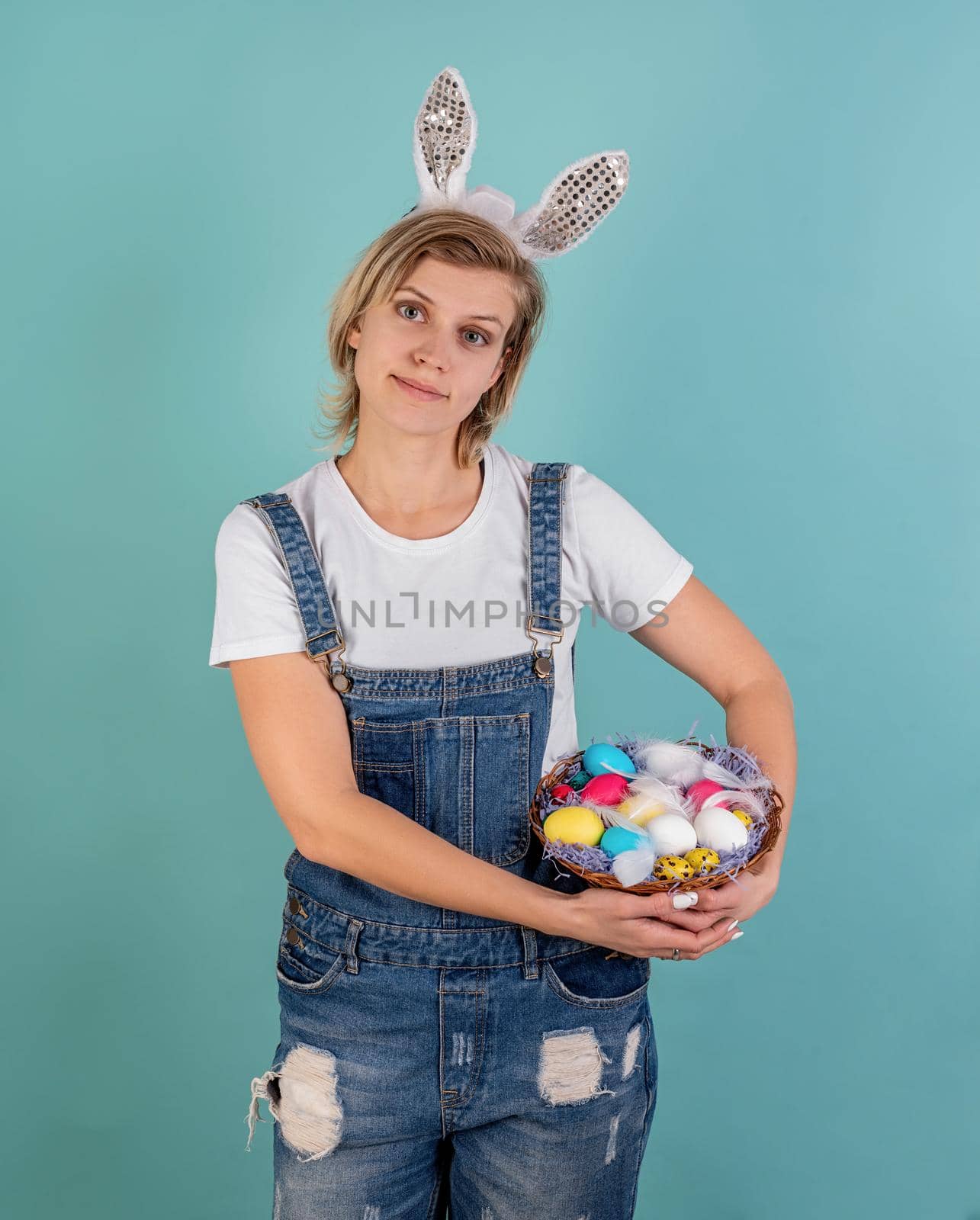 Easter holiday concept. Pretty young woman with bunny ears holding a basket with Easter colored eggs isolated on blue background