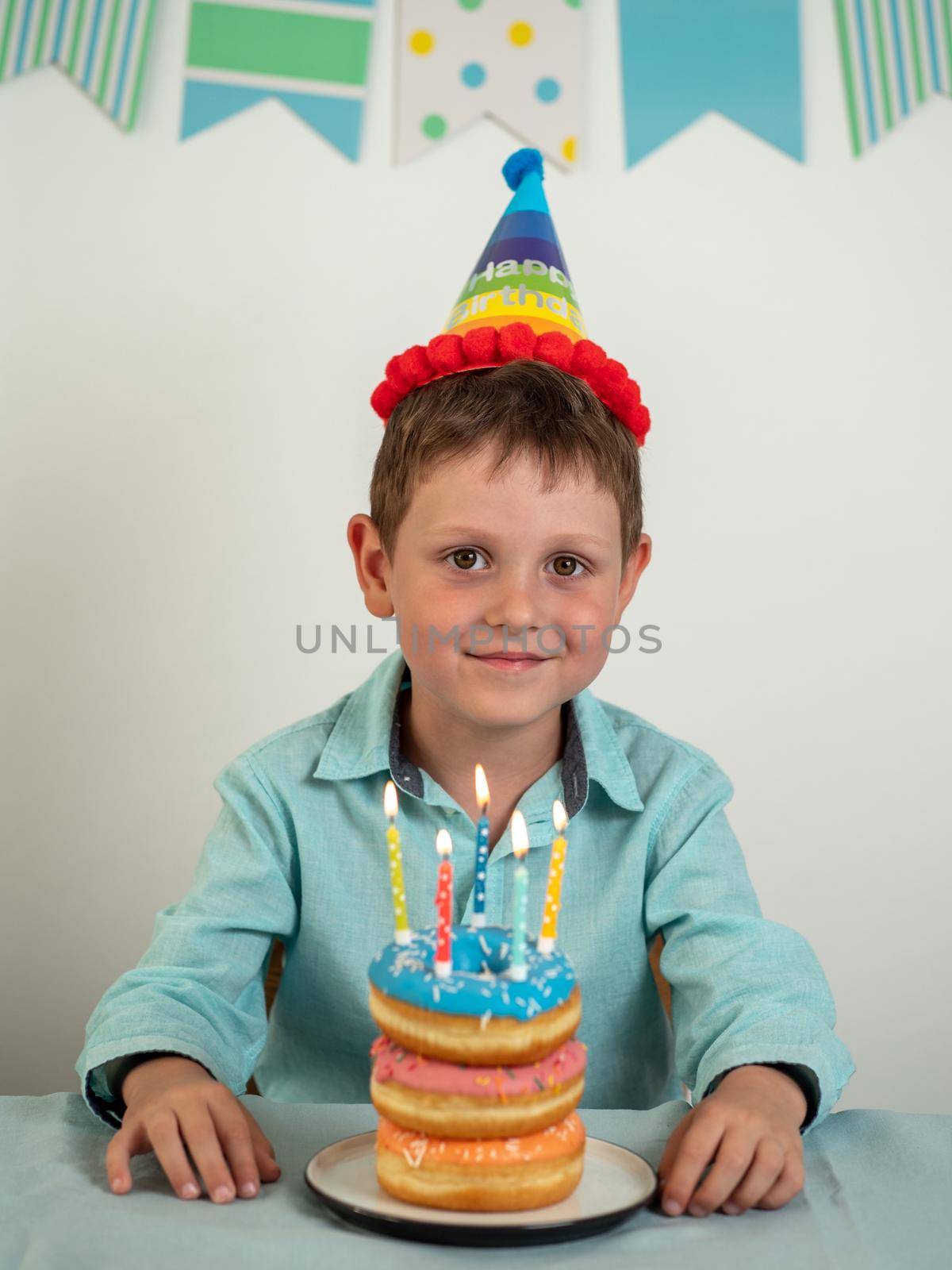 Five year old boy in his birthday and festie donuts cake with candles. Happy little child looking at camera and smiling. Plate with doughnut stack cake with candles on the table. Vertical.