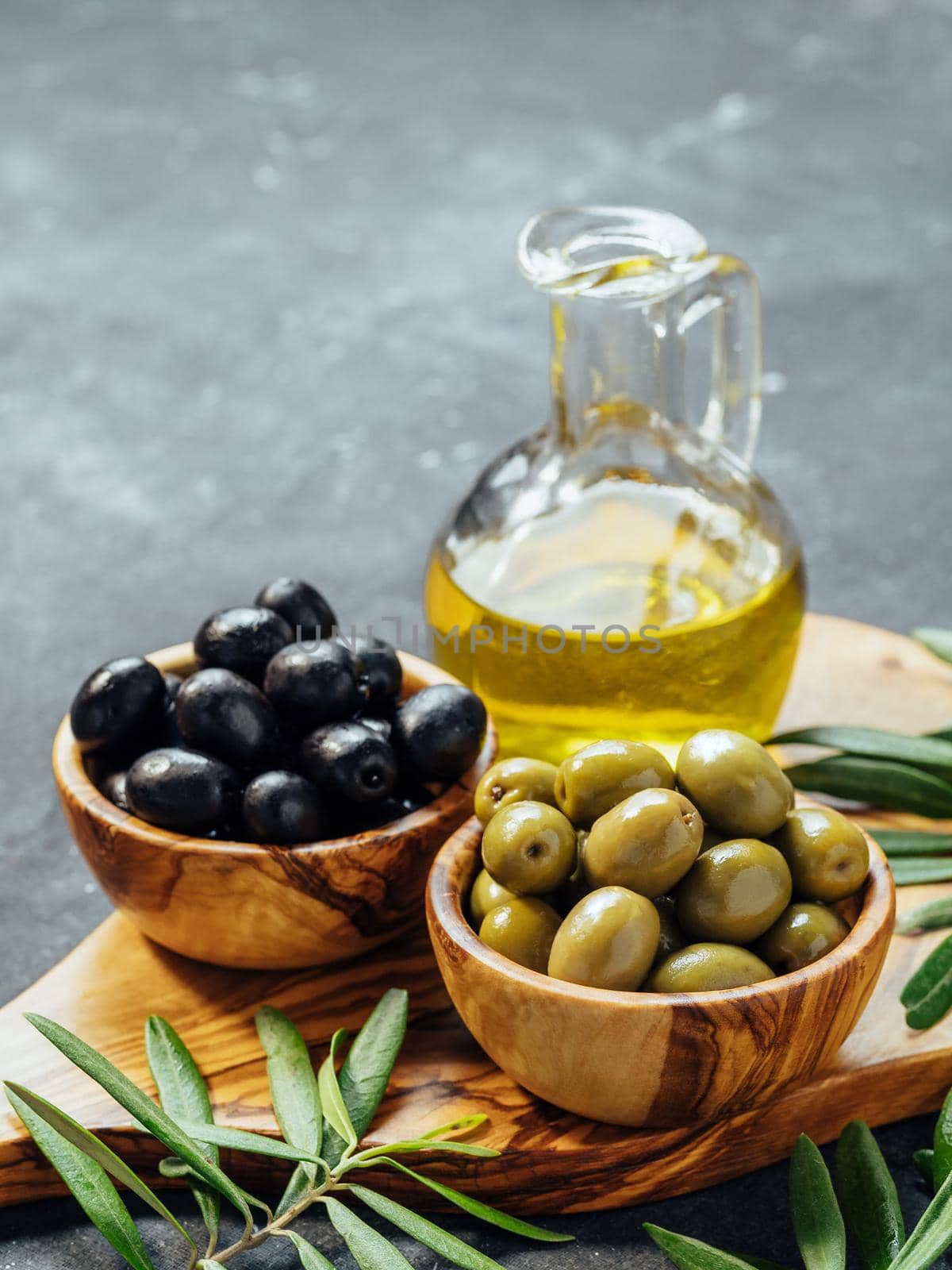 Set of green and black olives and olive oil on gray background. Different types of olives in olive wooden bowls and olive oil over wooden cutting board and fresh olive leaves. Copy space. Vertical