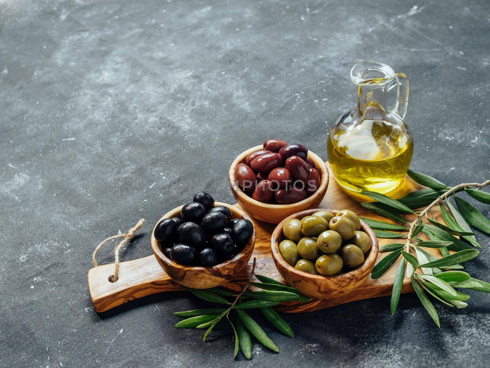 Set of green, black and red or pink olives and olive oil on gray background. Different types of olives in olive wooden bowls and olive oil over wooden cutting board. Copy space