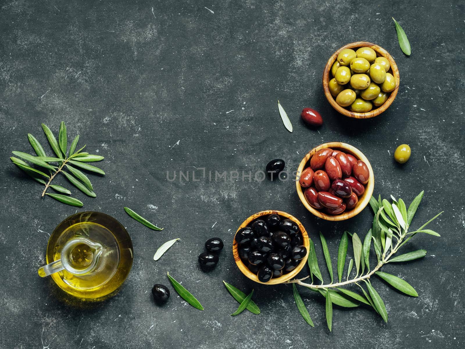 Set of green, black and red or pink olives and olive oil on dark background. Different types of olives in olive wooden bowls and olive oil over dark canvas background. Copy space. Top view or flat lay