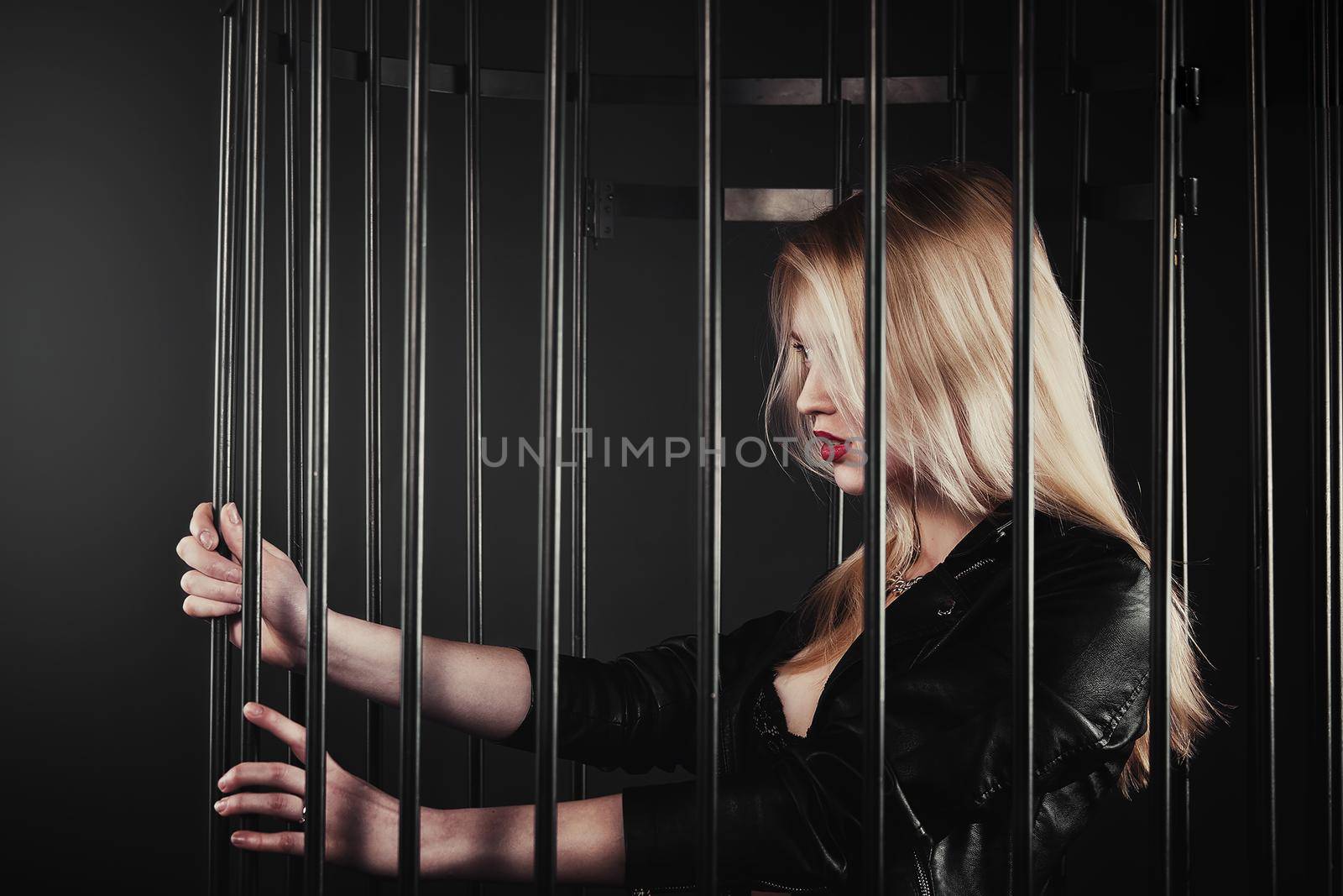 A beautiful, sexy young blond Girl in a black jacket is posing in an iron forged cage. The concept of domestic violence