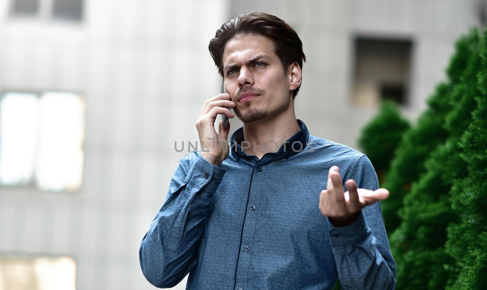 confident young man in full suit talking on the mobile phone and looking away while standing outdoors with cityscape in the background by Nickstock