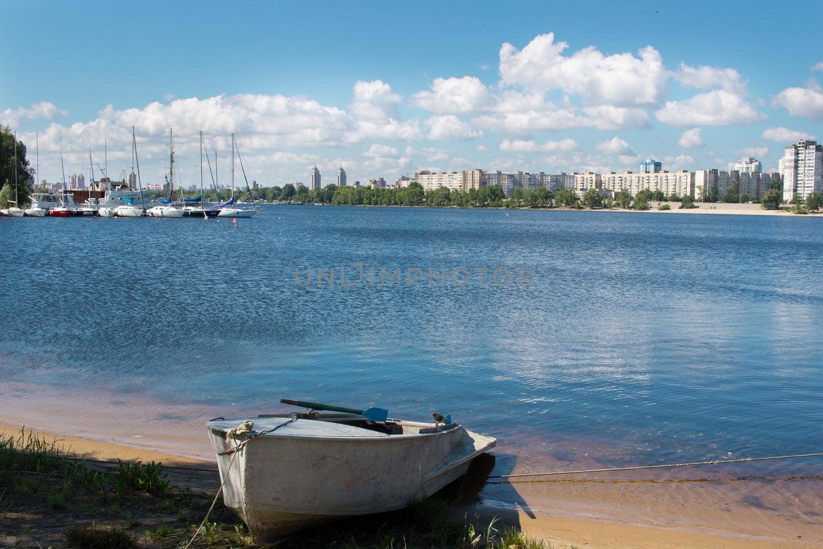 Small fishing boat with paddles stands near marina with sailing yachts and boats on river bank