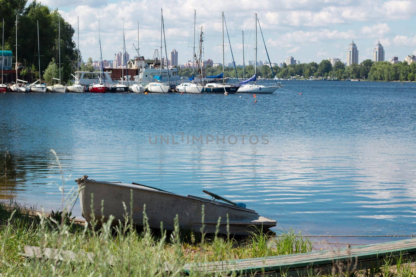 Small fishing boat with paddles stands near marina with sailing yachts on river bank