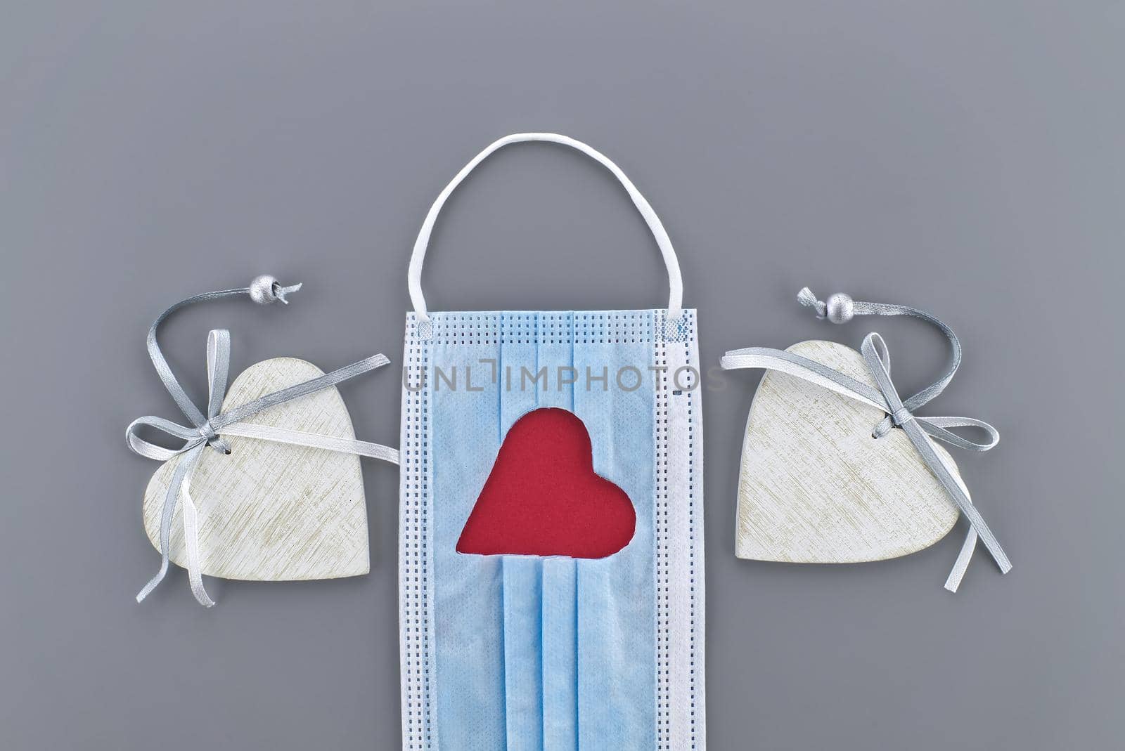 Two hearts between a medical face mask with cut out heart shape on a gray background. Stay at home, social distancing and quarantine concept