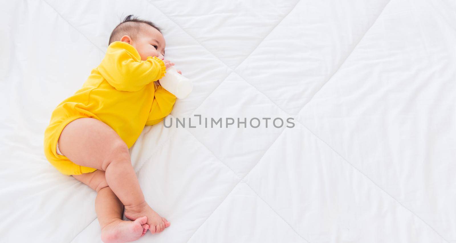 Asian beautiful little baby girl wearing a yellow dress eat milk sleeping feeding lying on the white bed, infant holding a bottle of milk, baby food concept