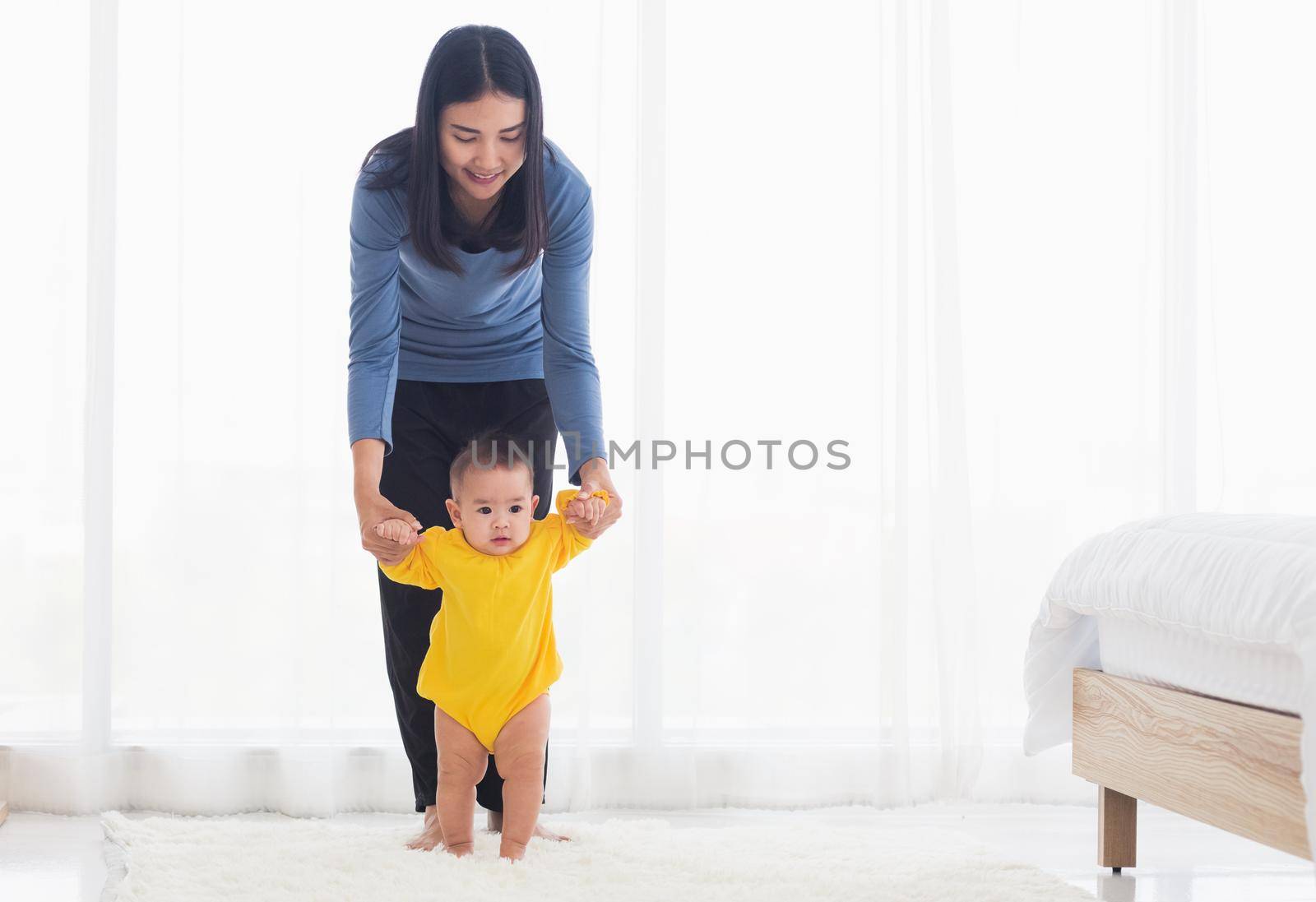 Asian little baby girl taking first steps learning to walk with mom help support the cute unstable walking toddler at home in bedroom. Happy family first steps parenthood concept