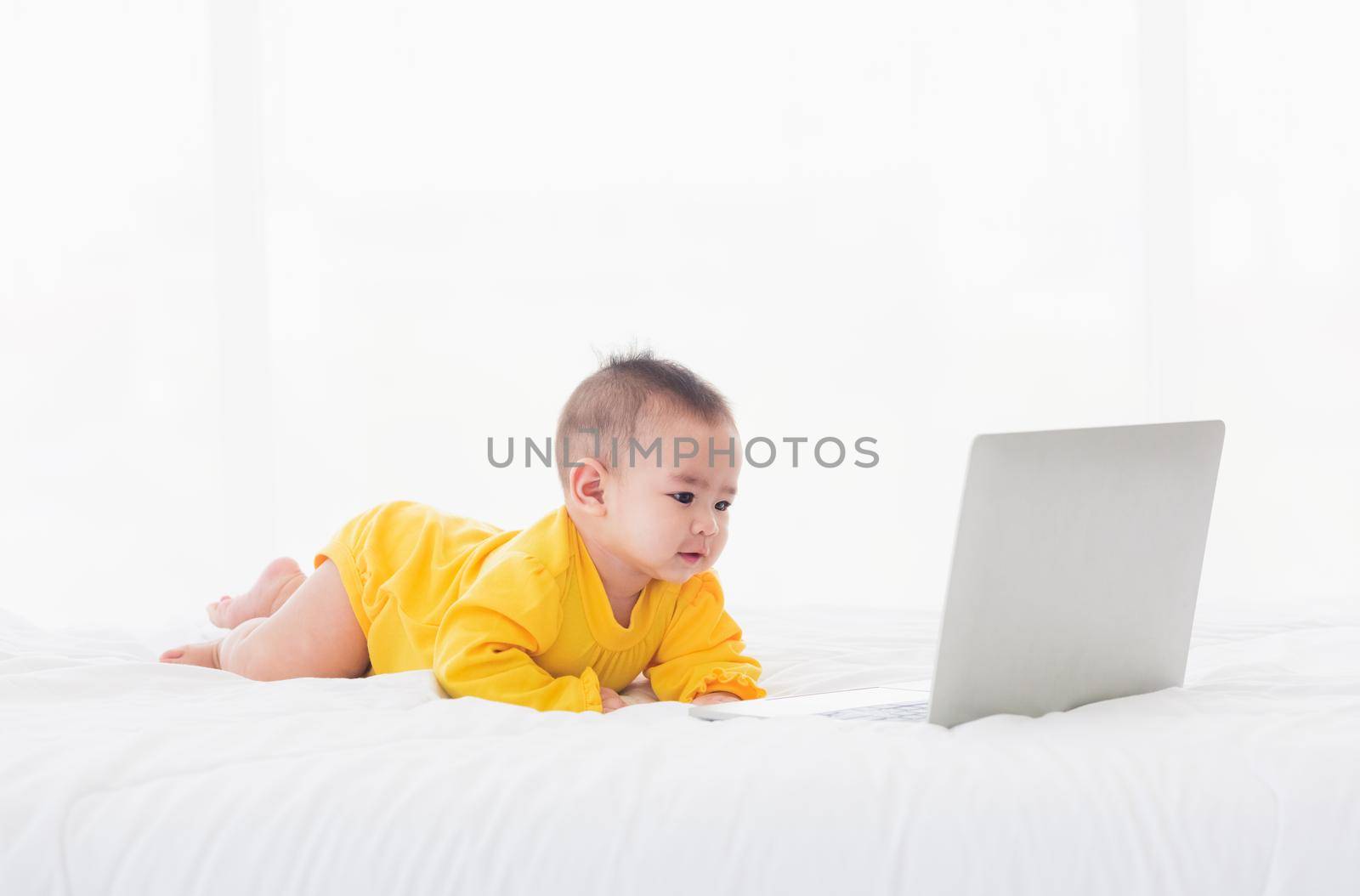 Asian little baby girl wearing a yellow dress typing hands on laptop computer keyboard and looking at monitor on the white bed. Little IT guy