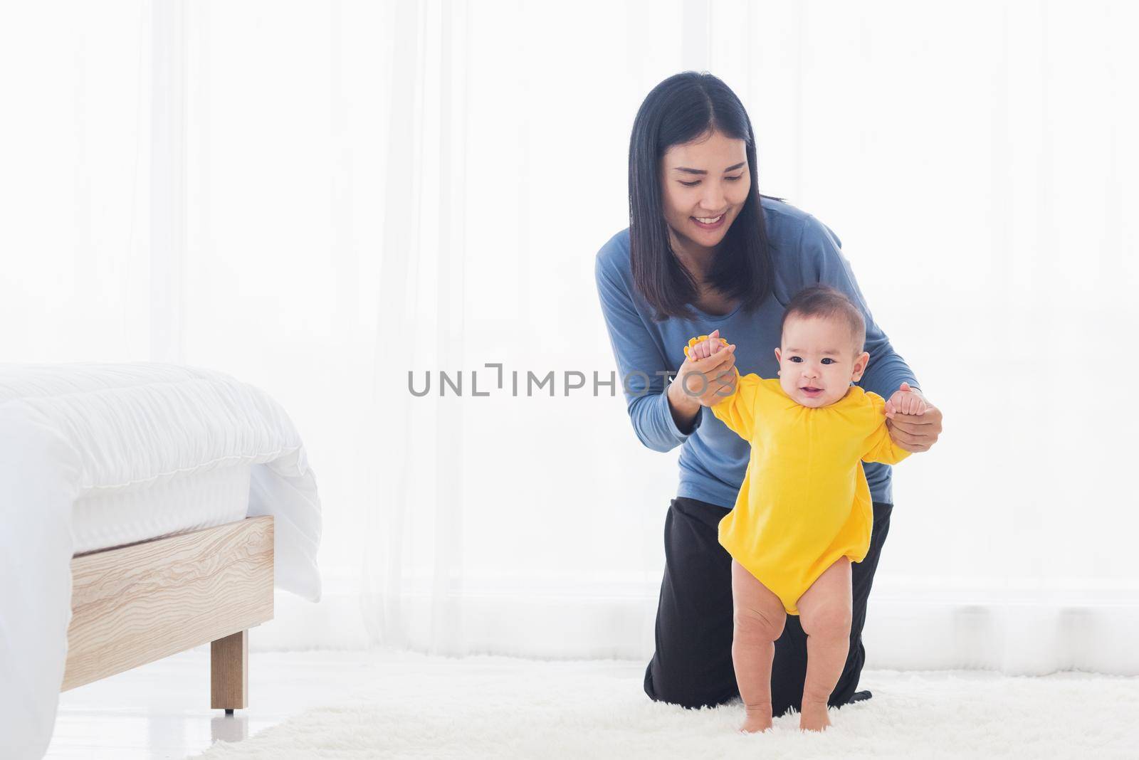 Asian little baby girl taking first steps learning to walk with mom help support the cute unstable walking toddler at home in bedroom. Happy family first steps parenthood concept