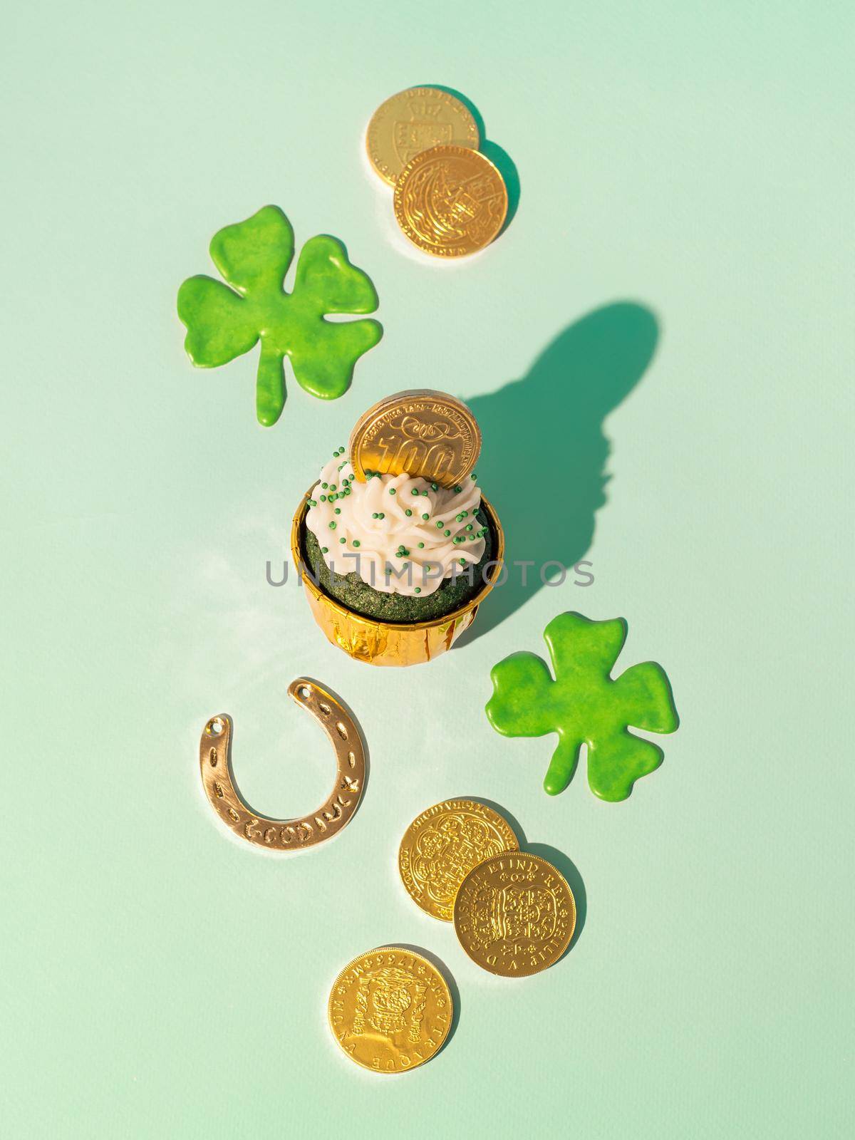 St Patrick's Day concept. Modern still life with lucky symbols and sweet food for Saint Patrick's Day party. Green velvet cupckake, shamrock, golden coins and horseshoe on green background. Vertical