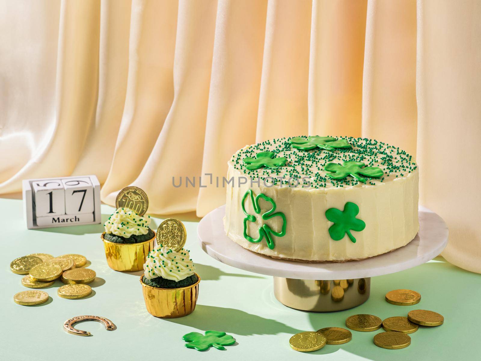 St Patrick's Day food concept. Modern still life with sweet food for Saint Patrick's Day party. Cake decorated shamrocks, green velvet cupckakes, golden coins and horseshoe on golden satin background