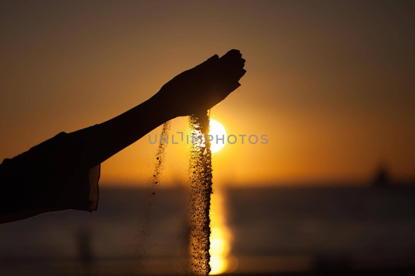 Silhouette of a girl's hand pouring sand from a beach against a yellow sky at sunset