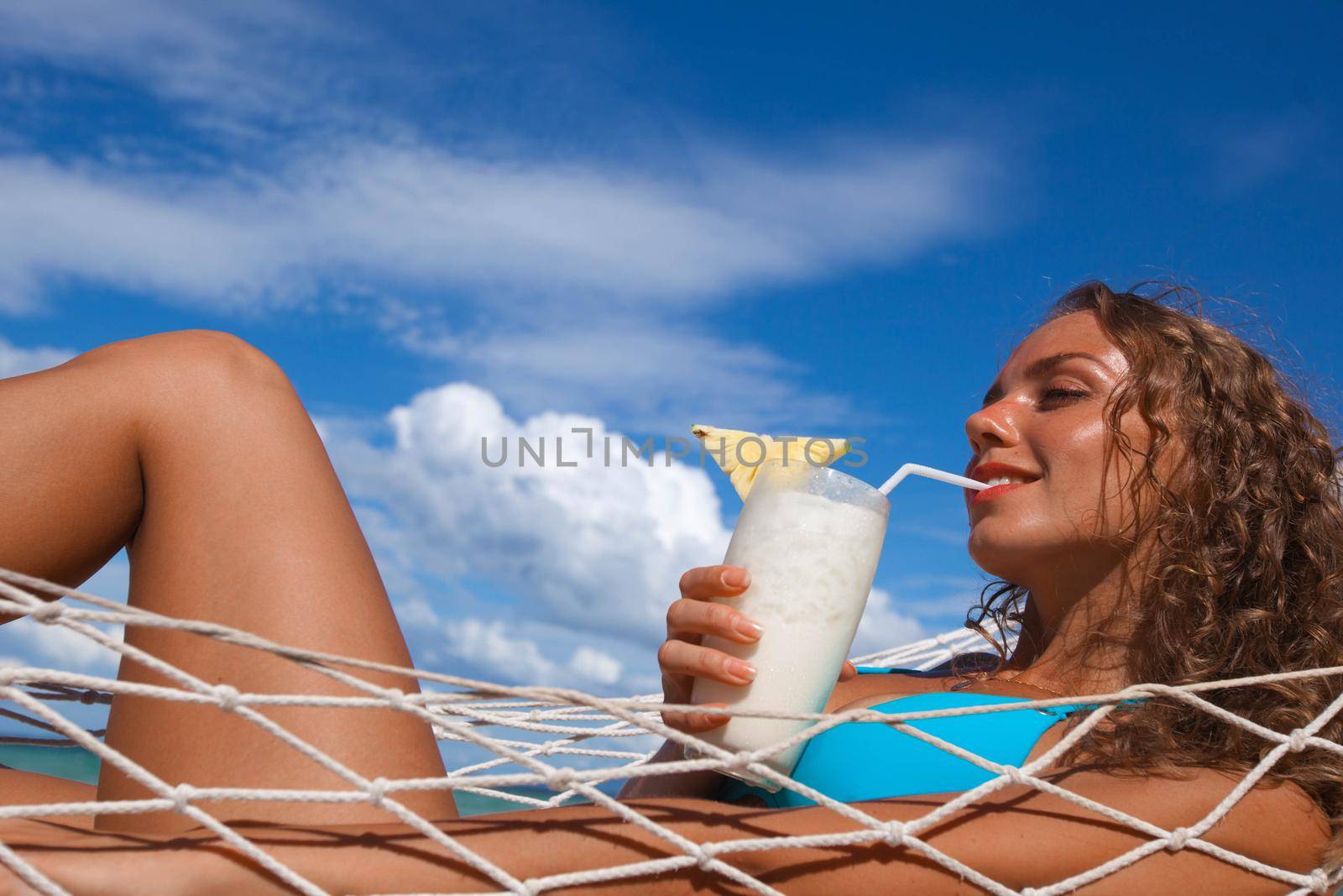 Pretty blonde girl in bikini sitting on hammock with Pina colada cocktail on the beach