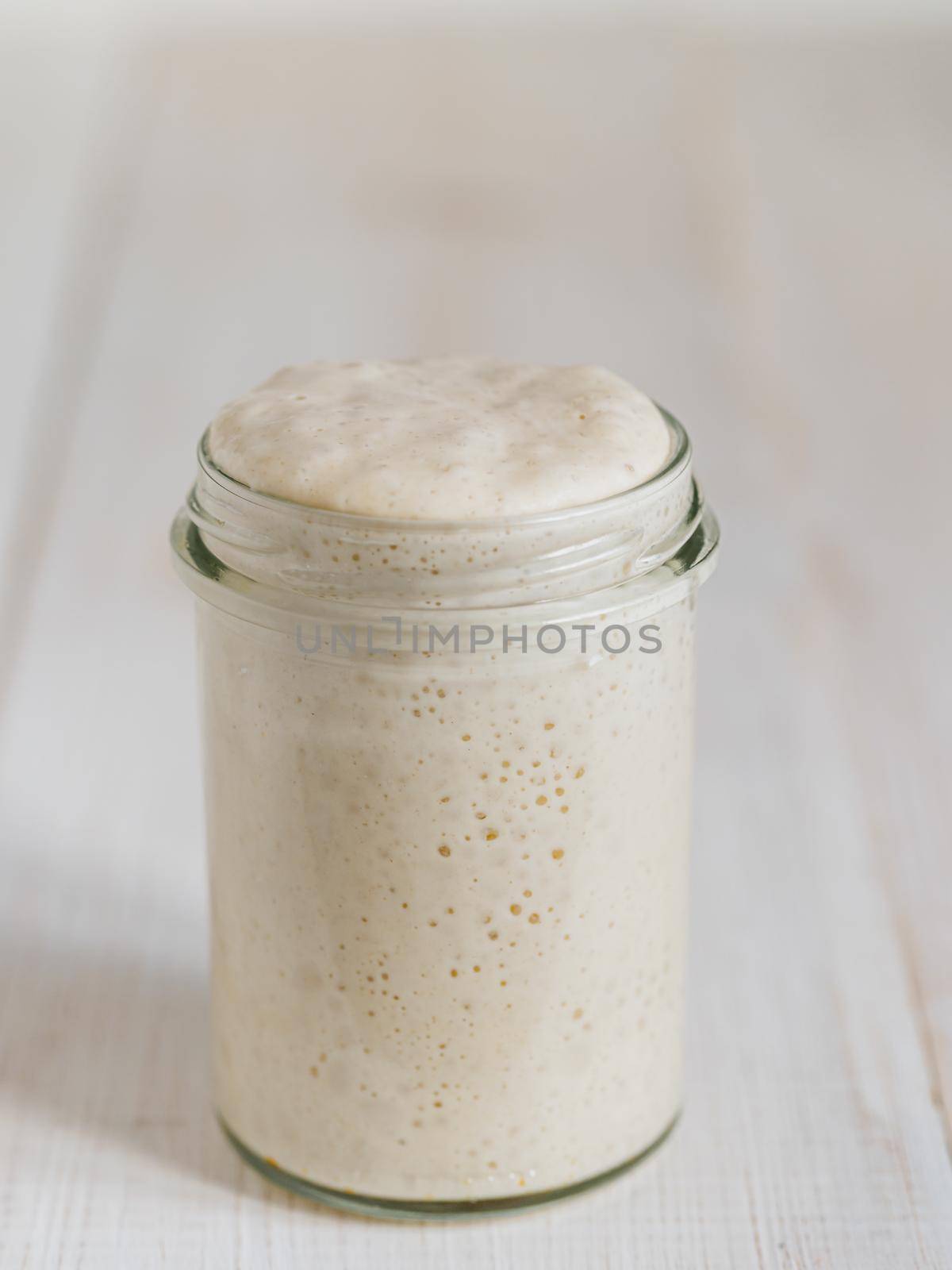 Wheat sourdough starter. Glass jar with sourdough starter on white wooden tabletop. Copy space for text or design. Vertical.