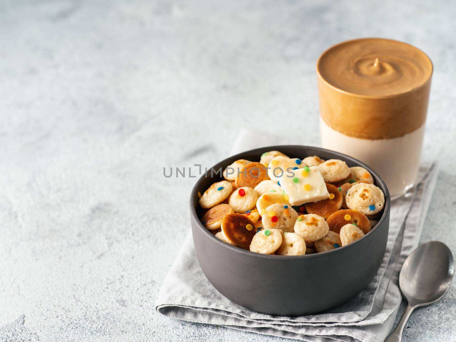 Mini pancakes cereal and dalgona coffee on gray cement background, copy space. Trendy food and drink - tiny panckakes served sprinkles and whipped instant coffee with milk or korean dalgona coffee